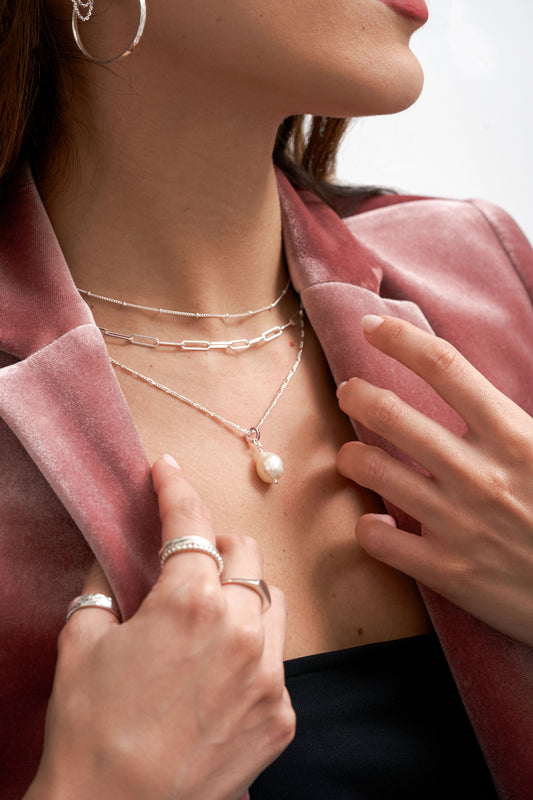 Woman wearing Single Freshwater Cultured pearl drop add-on charm, suspended from a figaro chain using L'ERA converter ring. Ring stack and necklace stack all in Sterling Silver. L'ERA Jewellery